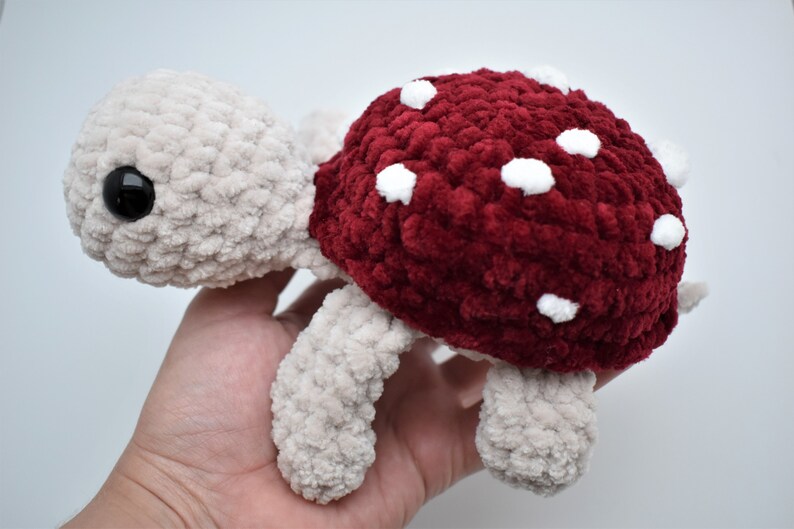 May include: A crocheted turtle toy with a mushroom-style shell. The turtle's body and limbs are beige, and the shell is a deep red with white spots. The turtle has a large, black eye, and is handmade.