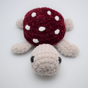 May include: A handmade plush turtle with a burgundy mushroom cap dotted with white spots. The turtle's body and limbs are a soft beige colour, and it has black eyes. This whimsical toy is perfect for children.