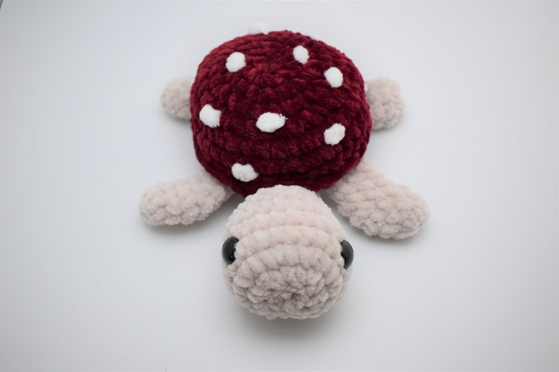May include: A handmade plush turtle with a burgundy mushroom cap dotted with white spots. The turtle's body and limbs are a soft beige colour, and it has black eyes. This whimsical toy is perfect for children.