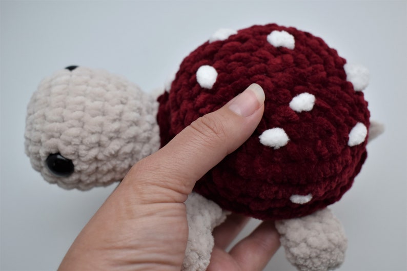 May include: A handmade plush turtle toy. The turtle has a burgundy shell with white spots and a light beige head, feet, and tail. The toy is made of a soft, textured material.