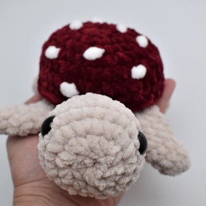May include: A handmade plush turtle with a mushroom cap. The turtle's body is a light beige colour, and the mushroom cap is a deep red with white spots. The turtle has black eyes and is made of soft, textured yarn.