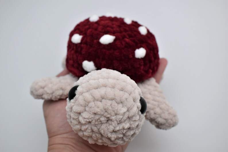 May include: A handmade plush turtle with a mushroom cap. The turtle's body is a light beige colour, and the mushroom cap is a deep red with white spots. The turtle has black eyes and is made of soft, textured yarn.