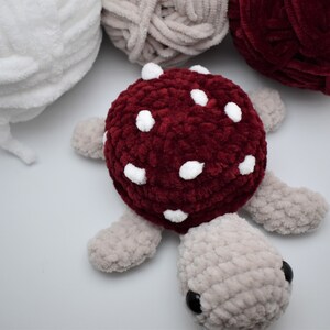 May include: A handmade plush turtle with a burgundy mushroom-shaped shell, decorated with white spots. The turtle's body and limbs are a soft grey, with black eyes. Several balls of yarn in white, grey, and burgundy are in the background.