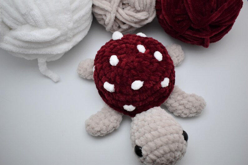 May include: A handmade plush turtle with a burgundy mushroom-shaped shell, decorated with white spots. The turtle's body and limbs are a soft grey, with black eyes. Several balls of yarn in white, grey, and burgundy are in the background.