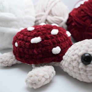 May include: A handmade crochet turtle with a burgundy shell and white spots. The turtle has a light grey head, feet, and a black eye. Soft yarn in white, beige, and burgundy are in the background.