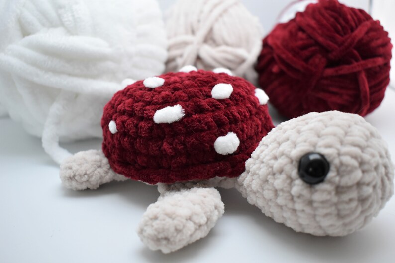 May include: A handmade crochet turtle with a burgundy shell and white spots. The turtle has a light grey head, feet, and a black eye. Soft yarn in white, beige, and burgundy are in the background.