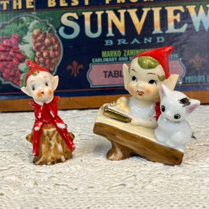 May include: Two ceramic figurines featuring elf-like characters. One elf sits on a brown tree stump wearing a red outfit. The second elf sits on a log next to a small white cat with a black spot, wearing a red hat and white shirt with gold accents.