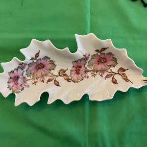 May include: A white porcelain leaf-shaped serving dish with a gold trim. The dish is decorated with a floral design of pink and brown flowers.