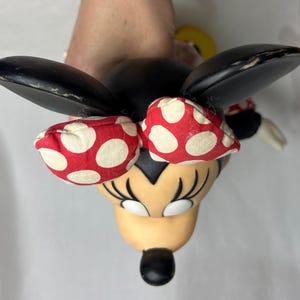 Peut inclure: Figurine de Minnie Mouse avec des oreilles noires, un n&oelig;ud &agrave; pois rouges et blancs et un visage bronz&eacute;. La figurine a des cils noirs et un nez noir. Les oreilles sont l&eacute;g&egrave;rement us&eacute;es.