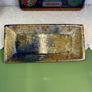 May include: A rectangular ceramic tray featuring a crystalline glaze with a gradient of deep blue, golden yellow, and brown tones. The surface displays intricate, star-like crystal formations scattered throughout the glossy, multi-colored finish.