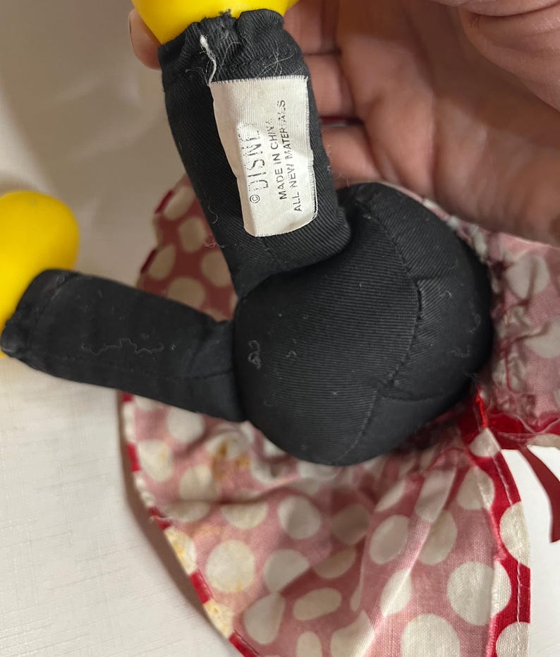 Peut inclure: Gros plan d'une poup&eacute;e en peluche Minnie Mouse. La poup&eacute;e a des jambes noires et un corps noir. La poup&eacute;e porte une jupe &agrave; pois rouges et blancs. L'&eacute;tiquette indique "DISNEY MADE IN CHINA ALL NEW MATERIALS."
