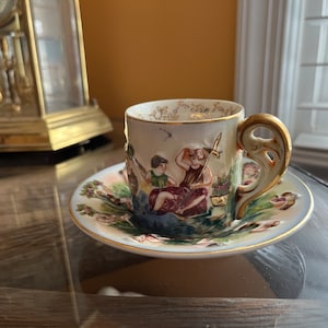 May include: A vintage porcelain teacup and saucer set with a hand-painted scene of two figures in a garden setting. The cup has a gold-trimmed handle and the saucer has a gold rim.