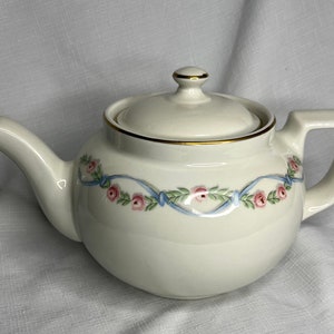 May include: Cream-colored ceramic teapot with a lid and handle. The teapot is decorated with a blue ribbon design, pink roses, and green leaves. The rim and the top of the lid are trimmed with gold.