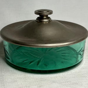 May include: A vintage green glass jar with a silver lid. The jar has an etched design and the lid has a knob on top.