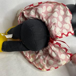Peut inclure: Une poup&eacute;e en peluche vintage de Minnie Mouse, avec des jambes et un corps en tissu noir, des chaussures jaunes et une robe &agrave; pois rouges et blancs. La robe de la poup&eacute;e a une bordure rouge et est l&eacute;g&egrave;rement froiss&eacute;e. La t&ecirc;te de la poup&eacute;e est noire.
