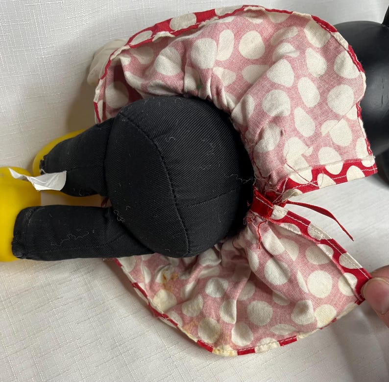Peut inclure: Une poup&eacute;e en peluche vintage de Minnie Mouse, avec des jambes et un corps en tissu noir, des chaussures jaunes et une robe &agrave; pois rouges et blancs. La robe de la poup&eacute;e a une bordure rouge et est l&eacute;g&egrave;rement froiss&eacute;e. La t&ecirc;te de la poup&eacute;e est noire.