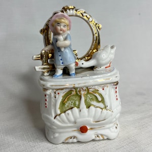 May include: A white porcelain trinket box with a decorative design, featuring a young girl in a blue coat and pink hat, holding a doll. A white goose and gold accents complete the scene. The box has a drawer with a red knob.