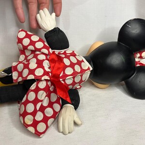 Peut inclure: Poup&eacute;e en peluche Minnie Mouse, avec une t&ecirc;te noire et de grandes oreilles, une robe &agrave; pois rouges et blancs et des gants blancs. La poup&eacute;e a des chaussures jaunes et un n&oelig;ud rouge. La poup&eacute;e est pos&eacute;e sur une surface blanche.