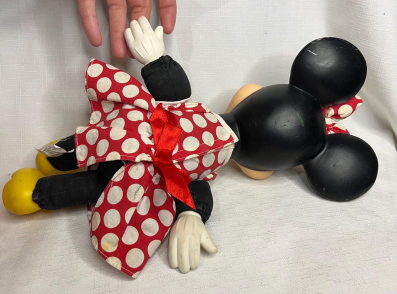 Peut inclure: Poup&eacute;e en peluche Minnie Mouse, avec une t&ecirc;te noire et de grandes oreilles, une robe &agrave; pois rouges et blancs et des gants blancs. La poup&eacute;e a des chaussures jaunes et un n&oelig;ud rouge. La poup&eacute;e est pos&eacute;e sur une surface blanche.
