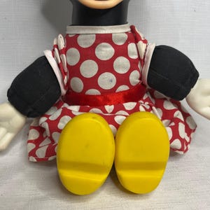 Peut inclure: Poup&eacute;e Minnie Mouse avec une t&ecirc;te et des bras noirs, des gants blancs et des pieds jaunes. Elle porte une robe rouge &agrave; pois blancs et un ruban rouge &agrave; la taille. Les bras de la poup&eacute;e sont tendus.