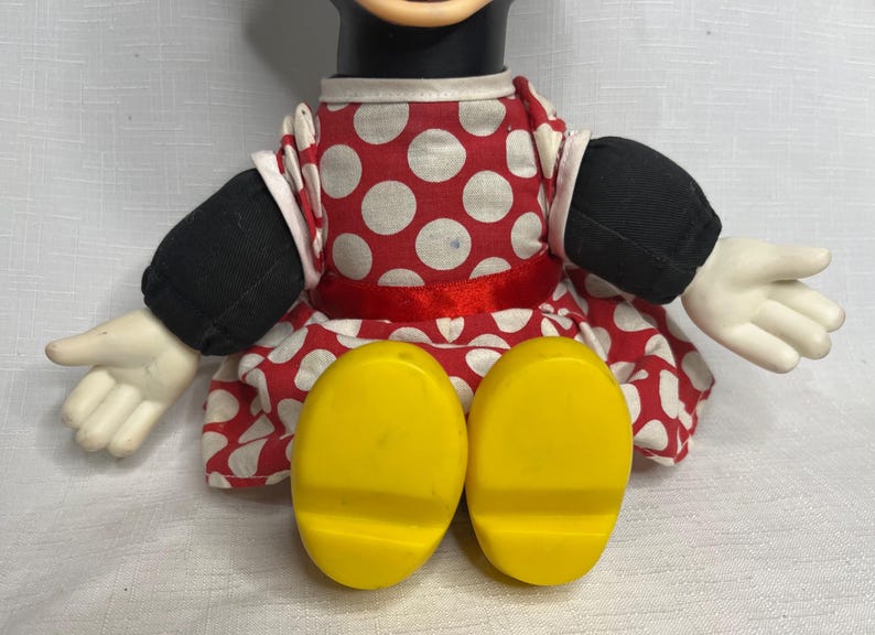 Peut inclure: Poup&eacute;e Minnie Mouse avec une t&ecirc;te et des bras noirs, des gants blancs et des pieds jaunes. Elle porte une robe rouge &agrave; pois blancs et un ruban rouge &agrave; la taille. Les bras de la poup&eacute;e sont tendus.