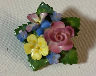 May include: A porcelain brooch featuring a cluster of colorful flowers, including a pink rose, yellow primrose, and blue forget-me-nots, set against green leaves.