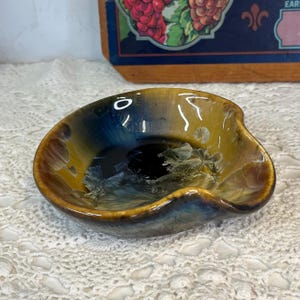 May include: A small, round, ceramic bowl with an irregular, wavy rim. The glaze features a blend of deep amber, golden yellow, and dark blue tones, with crystalline patterns radiating from the center of the bowl's interior surface.