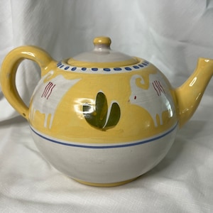 May include: A yellow ceramic teapot with a white and blue trim. The teapot has a white handle and spout and is decorated with two white goats and green leaves.