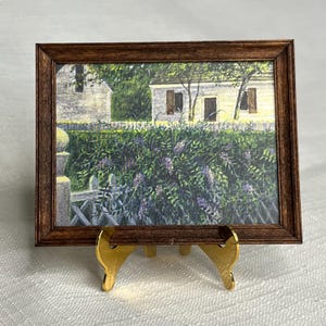 May include: Miniature framed print of a quaint country scene. The artwork depicts a white house with brown shutters, a white picket fence, and lush purple and green foliage. The frame is dark brown, and the print is displayed on a gold easel.