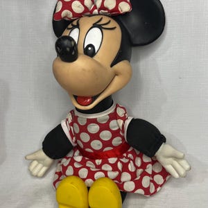 Peut inclure: Poup&eacute;e vintage de Minnie Mouse, v&ecirc;tue d'une robe et d'un n&oelig;ud &agrave; pois rouges et blancs. La poup&eacute;e a des oreilles noires, un nez noir et des chaussures jaunes. La poup&eacute;e est assise, les bras tendus.