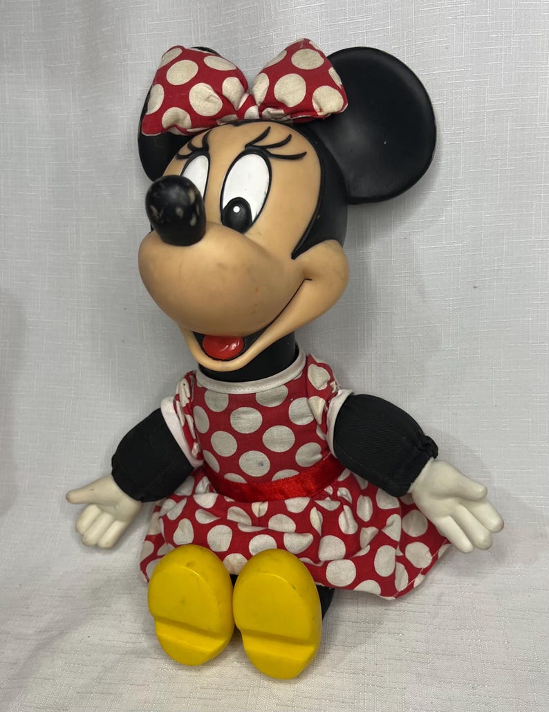 Peut inclure: Poup&eacute;e vintage de Minnie Mouse, v&ecirc;tue d'une robe et d'un n&oelig;ud &agrave; pois rouges et blancs. La poup&eacute;e a des oreilles noires, un nez noir et des chaussures jaunes. La poup&eacute;e est assise, les bras tendus.