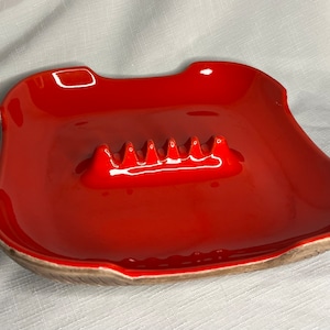 May include: Red ceramic ashtray with a scalloped edge and a raised, ridged center for holding cigarettes.