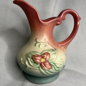 May include: A vintage ceramic pitcher with a curved spout and handle. The pitcher features a two-tone color scheme, with a pink top that fades into a cream-colored body. A floral design with a pink flower and green leaves decorates the front. The base is light blue.