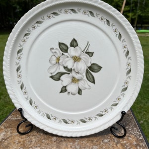 May include: White ceramic dinner plate with a floral design of white dogwood blossoms and green leaves. The plate has a rope-like edge and a border of small white flowers and green leaves.