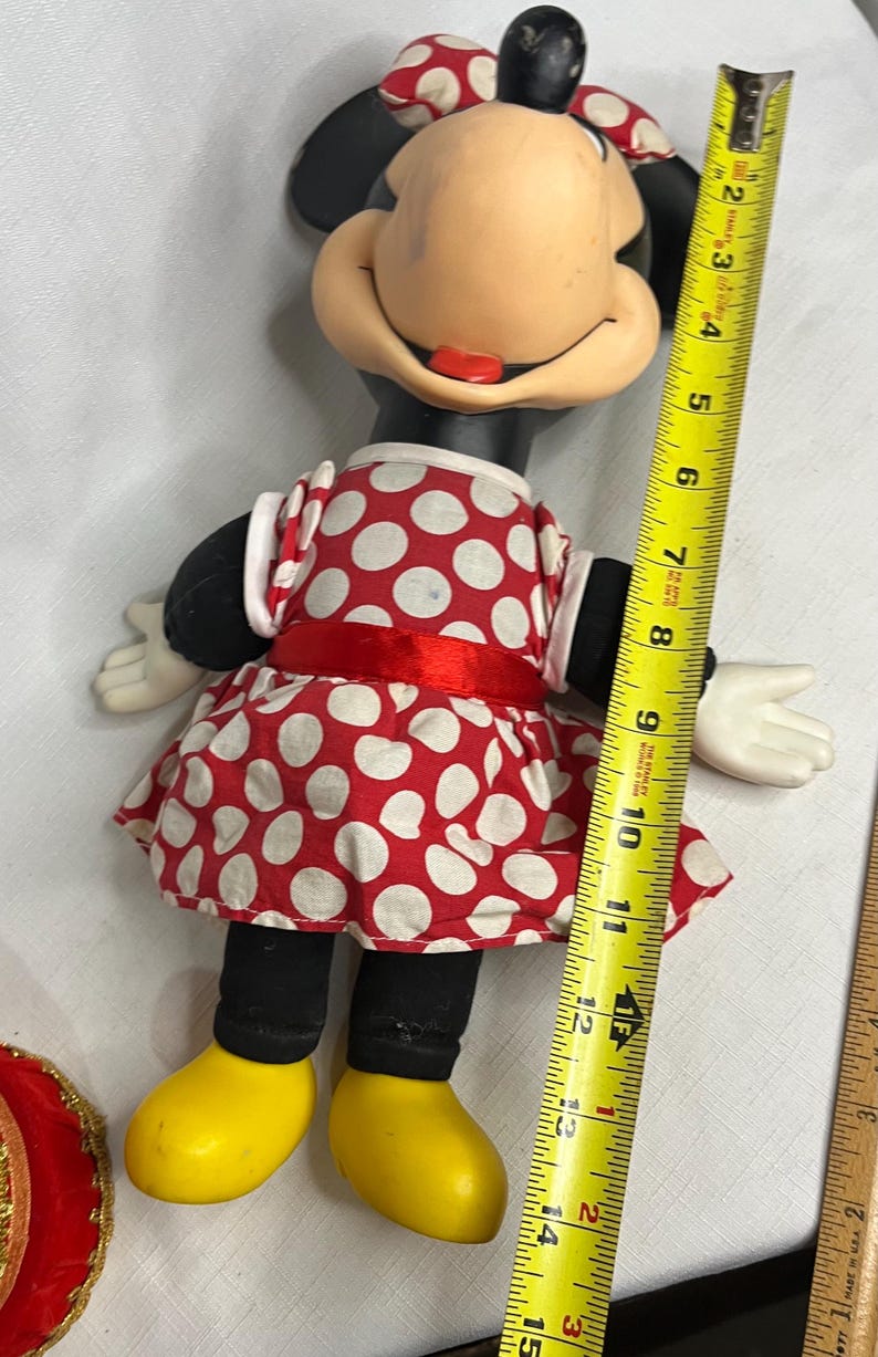 Peut inclure: Poup&eacute;e Minnie Mouse, environ 38 cm de haut, v&ecirc;tue d'une robe &agrave; pois rouges et blancs avec un n&oelig;ud assorti. La poup&eacute;e a des oreilles noires, des jambes noires, des chaussures jaunes et des gants blancs.