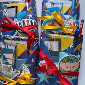 May include: Four gift-wrapped stationery sets. Each set is wrapped in blue paper with a star and lightning bolt pattern, tied with red or yellow ribbon. Pencils, pens, and stickers peek out from the top.