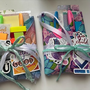 May include: Two colorful, handmade pen holders with pens, sticky notes, and stickers. The holders are decorated with floral and abstract patterns, tied with light blue and white ribbons. Text includes "God within her" and "I am a child".
