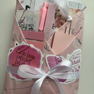 May include: A pink stationery set wrapped in patterned paper and tied with a white ribbon. The set includes a pen, highlighter, sticky notes, and decorative paperclips. The set has stickers with inspirational quotes.
