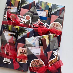 May include: Several gift packages wrapped in patterned paper and tied with red ribbons. Each package contains various items, including pens, cards, and stickers with motivational phrases like "Positivity is a choice" and "Never Give Up".