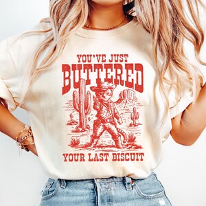 May include: A white t-shirt with a red graphic of a cat wearing a cowboy hat and holding a gun. The text "You've Just Buttered Your Last Biscuit" is printed below the cat.