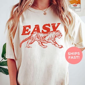 An ivory-colored shirt, illustrating an “EASY” text paired with a sleek tiger, blending minimal style with a hint of wild confidence — ideal for those who move through life with effortless strength.