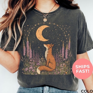 An pepper-colored shirt, illustrating a celestial fox nestled among flowers and gazing at the moon — good for nature lovers, stargazers, and those drawn to mystical woodland vibes.