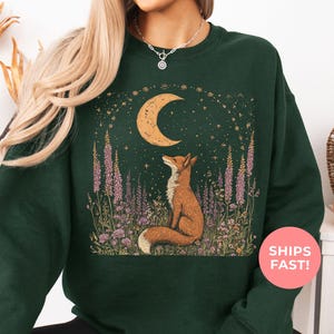 May include: Forest green sweatshirt featuring a fox gazing at a crescent moon and stars. The design includes purple flowers and a celestial theme. The text "SHIPS FAST!" is visible on the image. The color of the sweatshirt is forest green.