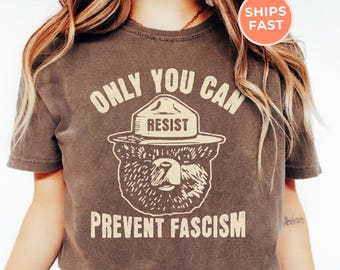 May include: An espresso-colored short-sleeve t-shirt featuring a graphic of a bear wearing a ranger hat. The text above the bear reads "ONLY YOU CAN RESIST" and the text below the bear reads "PREVENT FASCISM" in a light beige, distressed font.