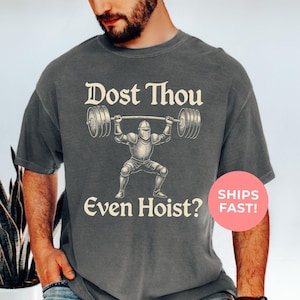 May include: Pepper-colored t-shirt featuring a knight lifting a barbell, with the text "Dost Thou Even Hoist?" in a vintage font. The design is a humorous take on medieval and fitness themes.
