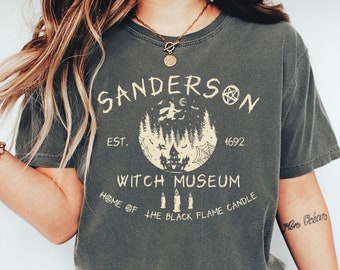 May include: A dark gray t-shirt with a vintage-style graphic design featuring the text "SANDERSON WITCH MUSEUM" with a witch flying on a broomstick, a house, and the text "EST. 1692" and "HOME OF THE BLACK FLAME CANDLE".