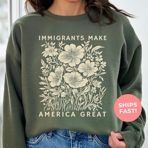 An militry green-colored sweatshirt, illustrating an “Immigrants Make America Great” design with soft pastel flowers—powerful yet gentle, blending activism with beauty to honor the strength, hope, and humanity of immigrant communities.