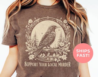 May include: Espresso-colored t-shirt featuring a detailed illustration of a raven perched on a branch within a circular frame of flowers and trees. The text "SUPPORT YOUR LOCAL MURDER" is printed below the image. The shirt is a vintage style.
