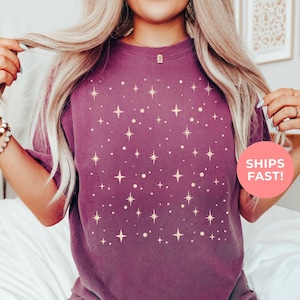 May include: Berry-colored t-shirt with a constellation design. The shirt features a pattern of stars and dots in a lighter shade. The text "SHIPS FAST!" is on a pink circle. The color is "BERRY".
