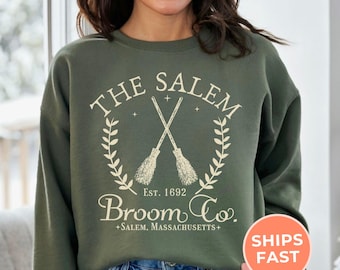 May include: Military green sweatshirt with the text "THE SALEM BROOM CO." and crossed broom graphics. The text includes "EST. 1692" and "+SALEM, MASSACHUSETTS+". The sweatshirt is a crew neck style.
